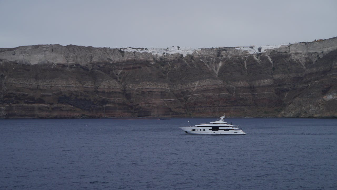 A luxury yacht cruises by the stunning cliffs of Santorini, Greece under a clear sky.